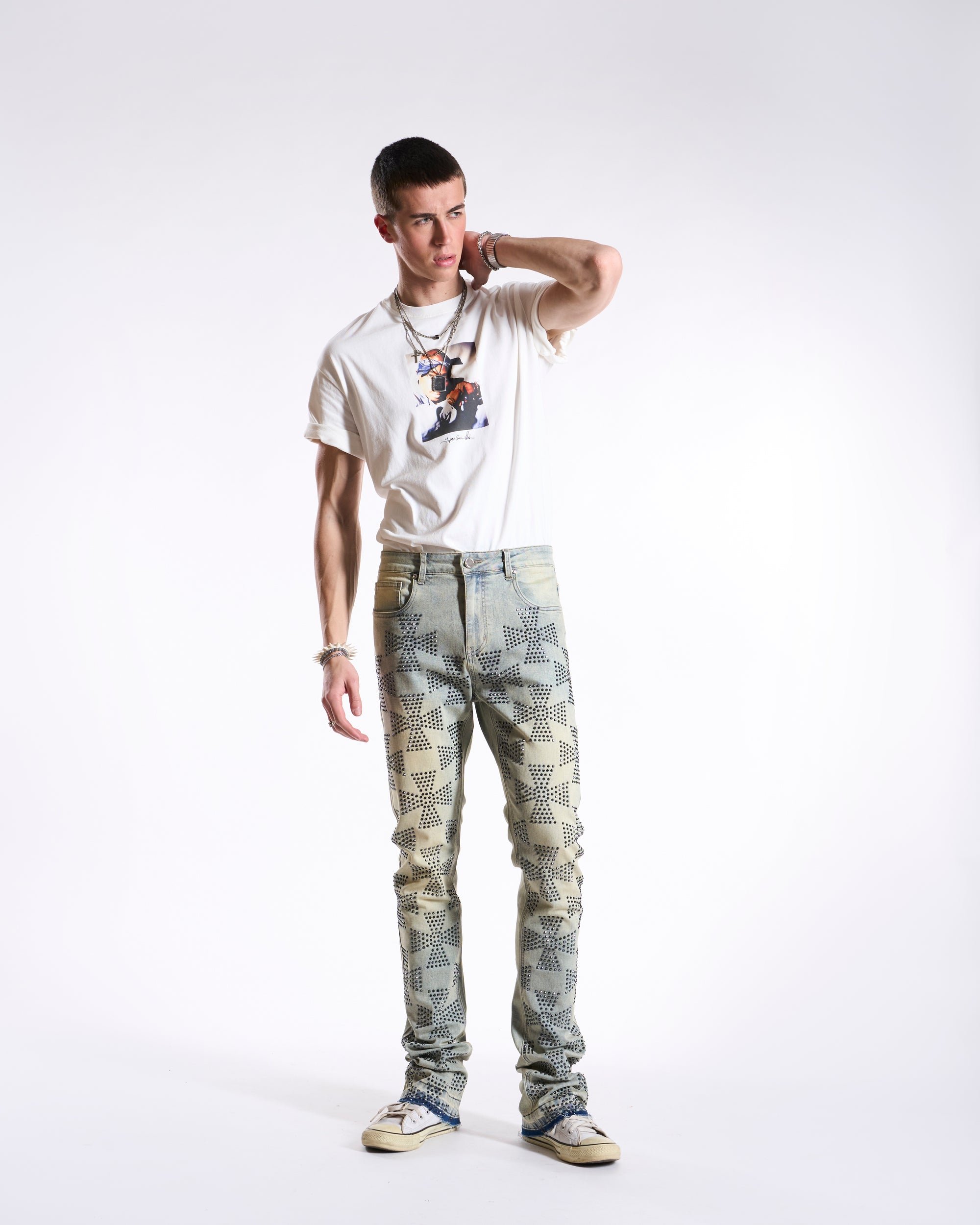 A man standing against a white background wearing camouflaged stacked jeans and a white t-shirt with a graphic on the front.