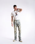 A man standing against a white background wearing camouflaged stacked jeans and a white t-shirt with a graphic on the front.